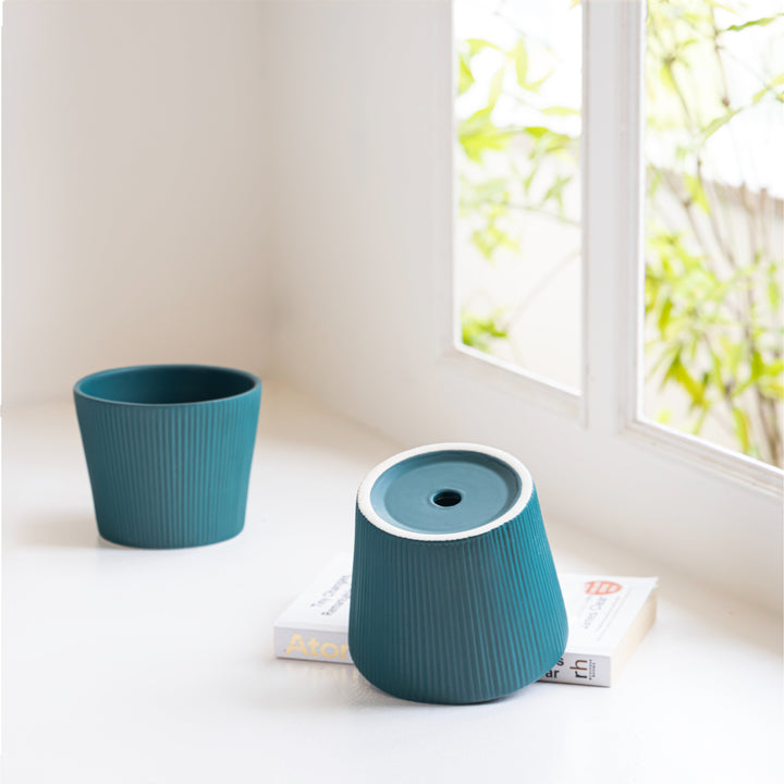 Teal ceramic planters placed near a bright window with a book in a minimalist decor setting.