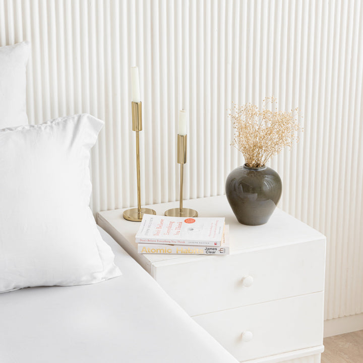 Minimalist bedroom setup featuring a white bedside table with books, brass candle holders, and a The Elegant Vase holding dried flowers next to a white pillow and bed.