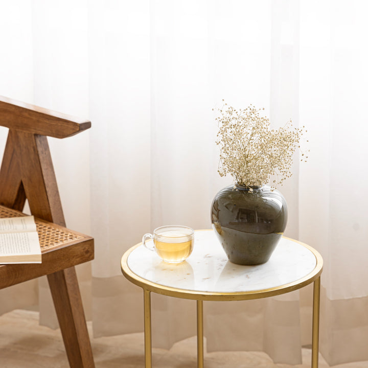 Cozy scene with a ceramic vase filled with dried flowers on a marble side table, next to a glass cup of tea and a wooden chair, against a soft white curtain background.